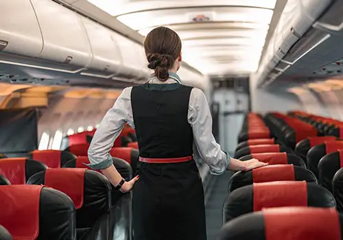 A flight attendant with her back to the camera stands in the aisle of an empty single aisle plane.