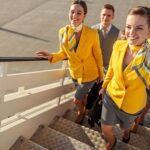Smiling flight attendants wearing yellow and gray walking up a flight of stairs.
