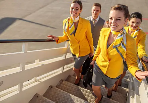Smiling flight attendants wearing yellow and gray walking up a flight of stairs. 
