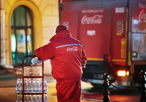 A Coca Cola delivery driver dressed for cold weather is leaning on a dolly loaded with cases of bottled drinks.