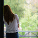 An introverted young woman standing in a doorway looking outside at trees and nature.
